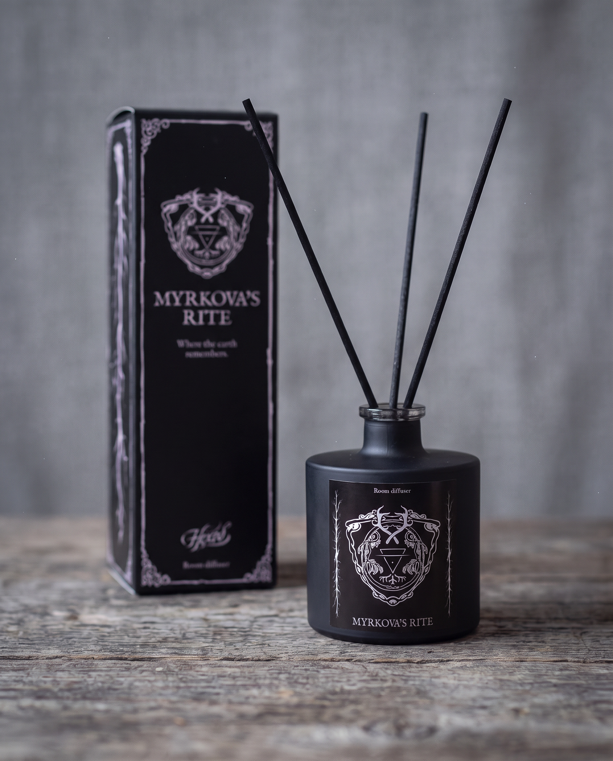 Myrkova's Rite room diffuser by HEXED, a matte black glass bottle with black reeds and a silver crest label, with its black illustrated gift box softly out of focus behind it on a weathered wooden surface