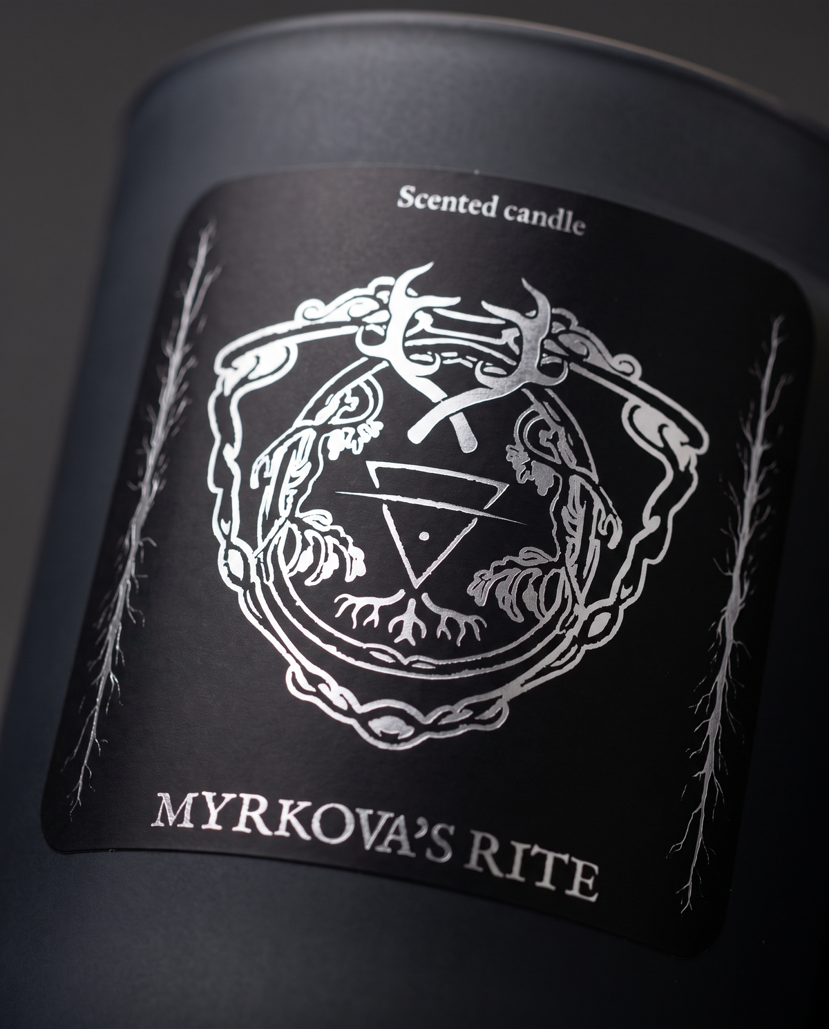 Close-up of the silver-printed Myrkova's Rite label on the matte black HEXED scented candle jar, showing the antler and root crest