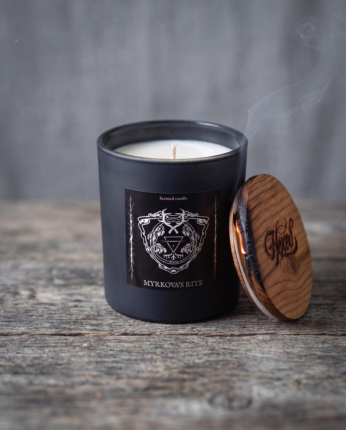 Myrkova's Rite scented candle by HEXED in a matte black glass jar with a silver crest label, its oak lid with a charred edge leaning against the jar on a weathered wooden surface