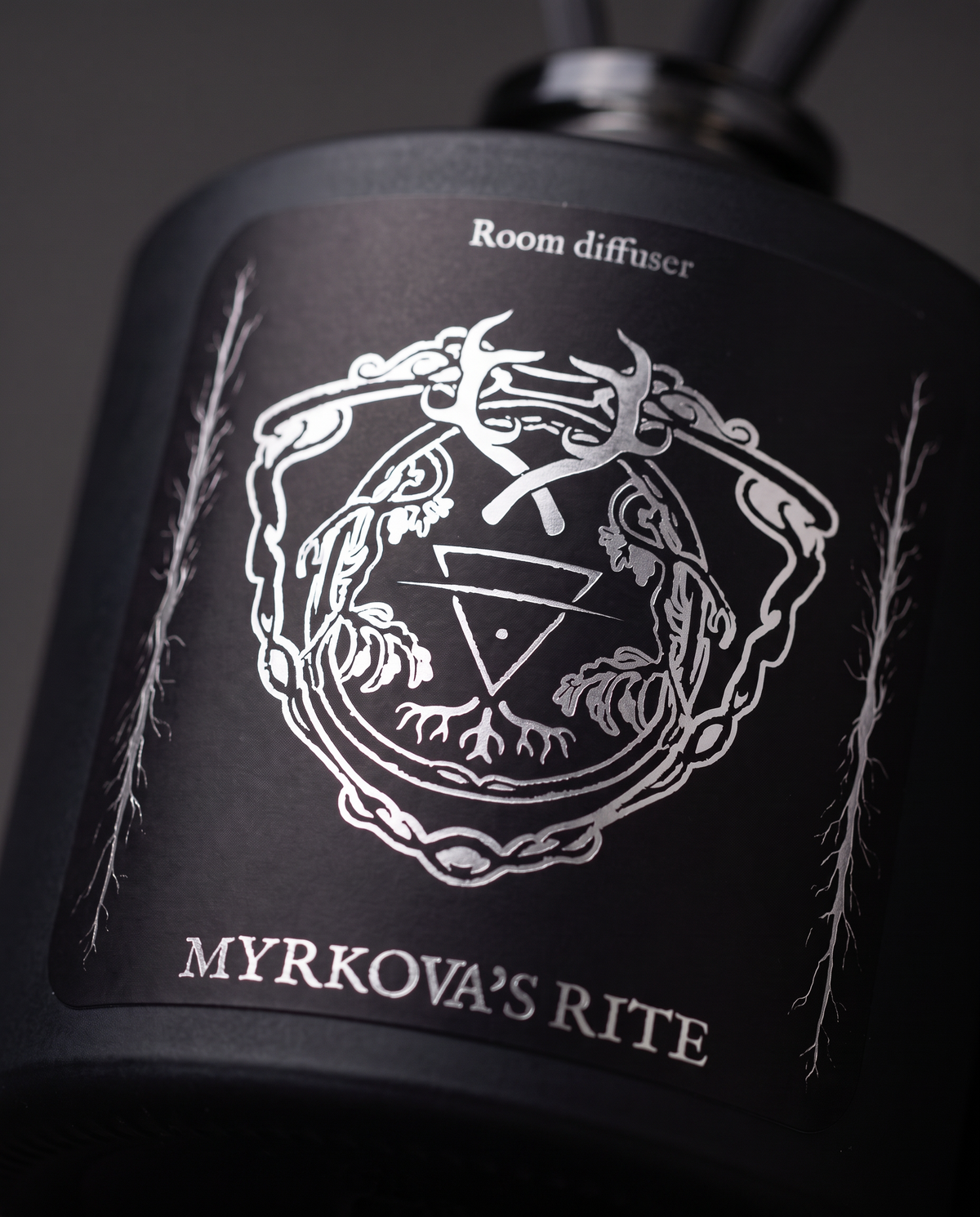 Close-up of the silver-printed Myrkova's Rite label on the matte black HEXED room diffuser bottle, showing the antler and root crest